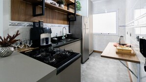 A modern kitchen with a gas stove, stainless steel refrigerator, wooden shelves, and a small dining table. - Apto a 500m da Praia Canto do Forte - Praia Grande (Praia Grande)