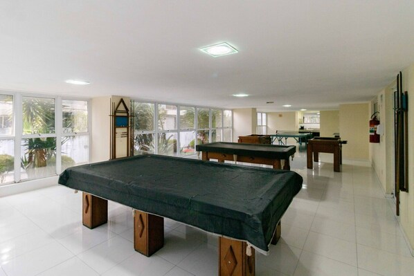 A spacious indoor area with a pool table, another table, and a billiards table. Large windows allow natural light and views of the outdoors.