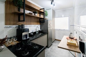 A compact kitchen with a black stove, stainless steel refrigerator, and wooden shelves. - Apto a 500m da Praia Canto do Forte - Praia Grande (Praia Grande)
