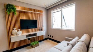 A modern living room with a flat-screen TV mounted on a wooden panel wall, a beige sofa, and a large window providing natural light. - Flat Equipado em Tima Localiza o Wi-fi e Lazer (Barueri)