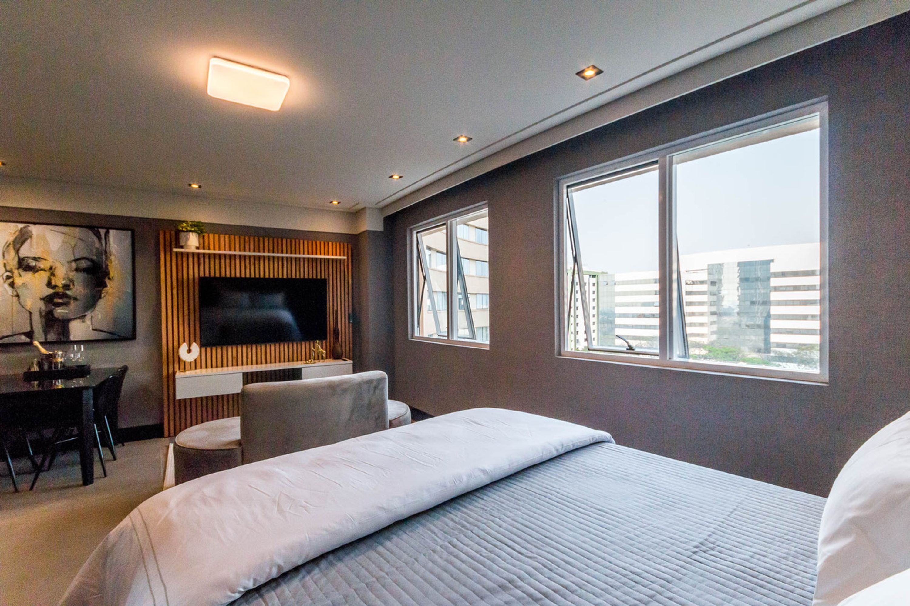 A modern hotel room with a large bed, a TV mounted on a wooden panel, a dining area with a table and chairs, and large windows offering a view of buildings.