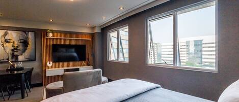 A modern hotel room with a large bed, a TV mounted on a wooden panel, a dining area with a table and chairs, and large windows offering a view of buildings.