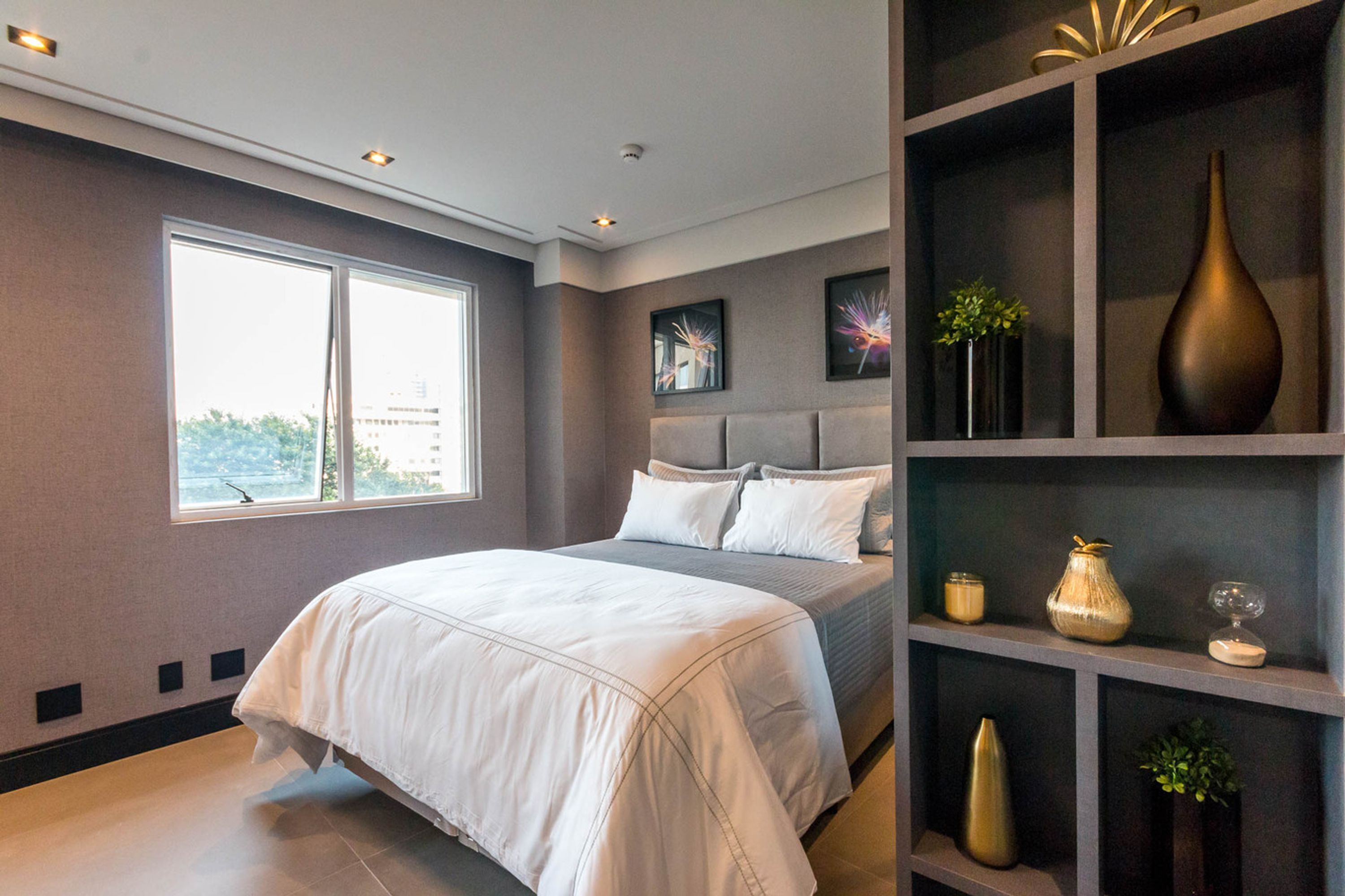 Un dormitorio moderno con una cama grande, cabecera, una ventana y una estantería con objetos decorativos.