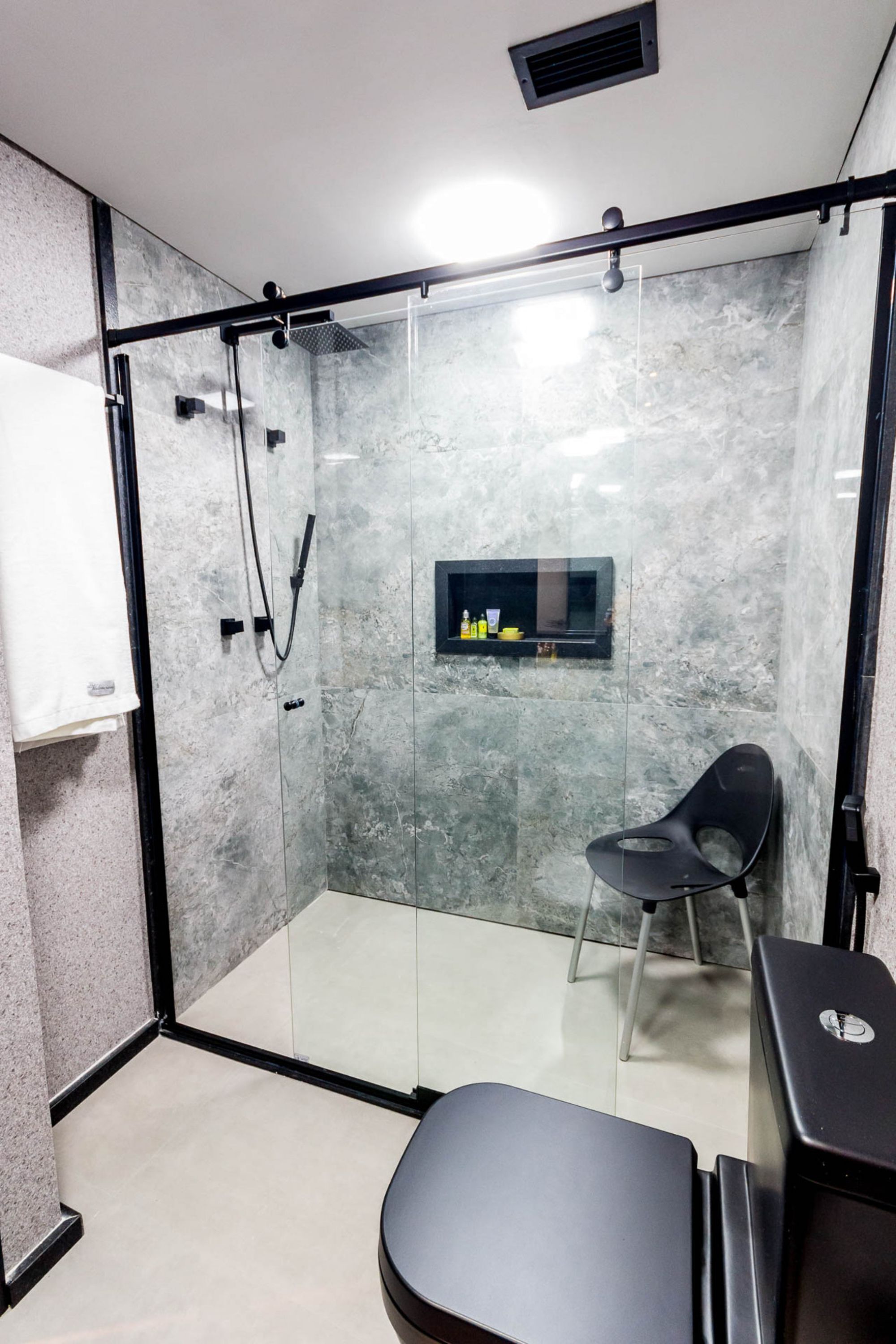 A modern bathroom with a glass shower enclosure, a black chair, and a small shelf with toiletries.