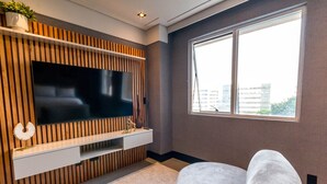 Un salon moderne avec un téléviseur à écran plat fixé sur un mur recouvert de panneaux en bois, un canapé blanc et une grande fenêtre donnant sur la ville.