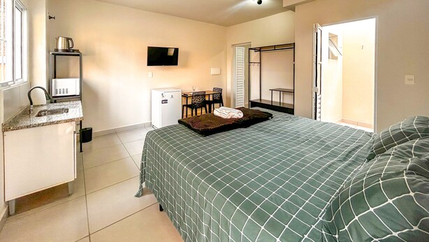 Quarto de hotel com uma cama, uma escrivaninha com computador, televisão, cozinha compacta e um guarda-roupa.