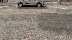 Um carro estacionado em frente a um prédio com uma placa de número 109, uma cerca de arame farpado e um poste de eletricidade com cabos.