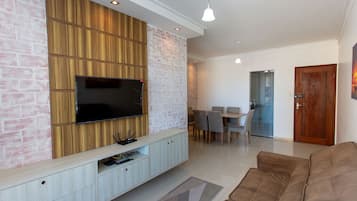 A modern living room with a flat-screen TV mounted on a wooden panel wall, a brown sofa, and a dining area with chairs and a table.