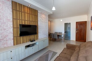 A modern living room with a flat-screen TV mounted on a wooden panel wall, a brown sofa, and a dining area with chairs and a table. - Apto com Ar-condicionado a 350m do mar em Aracaju (Aracaju)