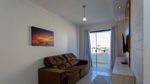 A modern living room with a brown sofa, a painting of a sunset, a TV mounted on the wall, and a balcony with curtains. - Apto com Ar-condicionado a 350m do mar em Aracaju (Aracaju)