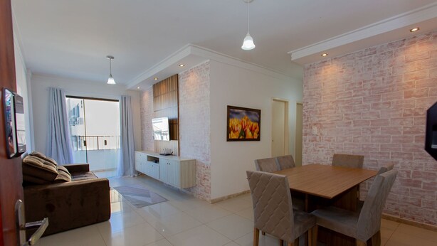 A modern living room with a sofa, a dining table with chairs, a kitchen area, and a large window with curtains. - Apto com Ar-condicionado a 350m do mar em Aracaju (Aracaju)