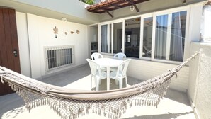 A hammock in a sunlit outdoor area with a white table and chairs, a window with curtains, and a wall-mounted signboard. - Aconchegante e Espa oso Apto c Piscina e Churrasq (Cabo Frio)