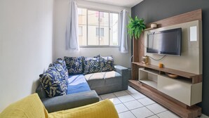 A modern living room with a sofa, a television mounted on the wall, and a window with curtains. - Flat Confort vel com Wi-fi em Santa Cruz RJ (Rio de Janeiro)