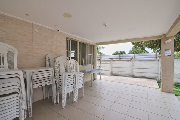 A covered patio area with stacked plastic chairs and a table, a tiled floor, and a view of a fence and greenery outside.