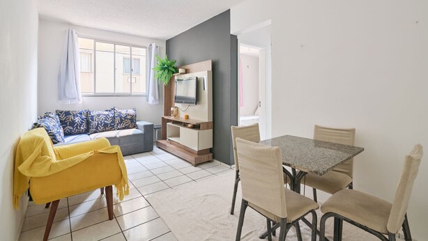 A modern living room with a sofa, a yellow chair, a dining table with chairs, a TV on a wooden stand, and a window with curtains. - Flat Confort vel com Wi-fi em Santa Cruz RJ (Rio de Janeiro)