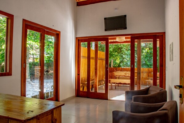 Una habitación amplia con una mesa de madera, un sofá y una puerta corrediza de vidrio que da a un área exterior con terraza de madera y sillas.