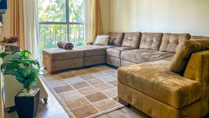 A living room with a sectional sofa, a small ottoman, a potted plant, and a large window with curtains. - Apto Espa oso c Wifi pr x a Marginal Pinheiros SP (São Paulo)