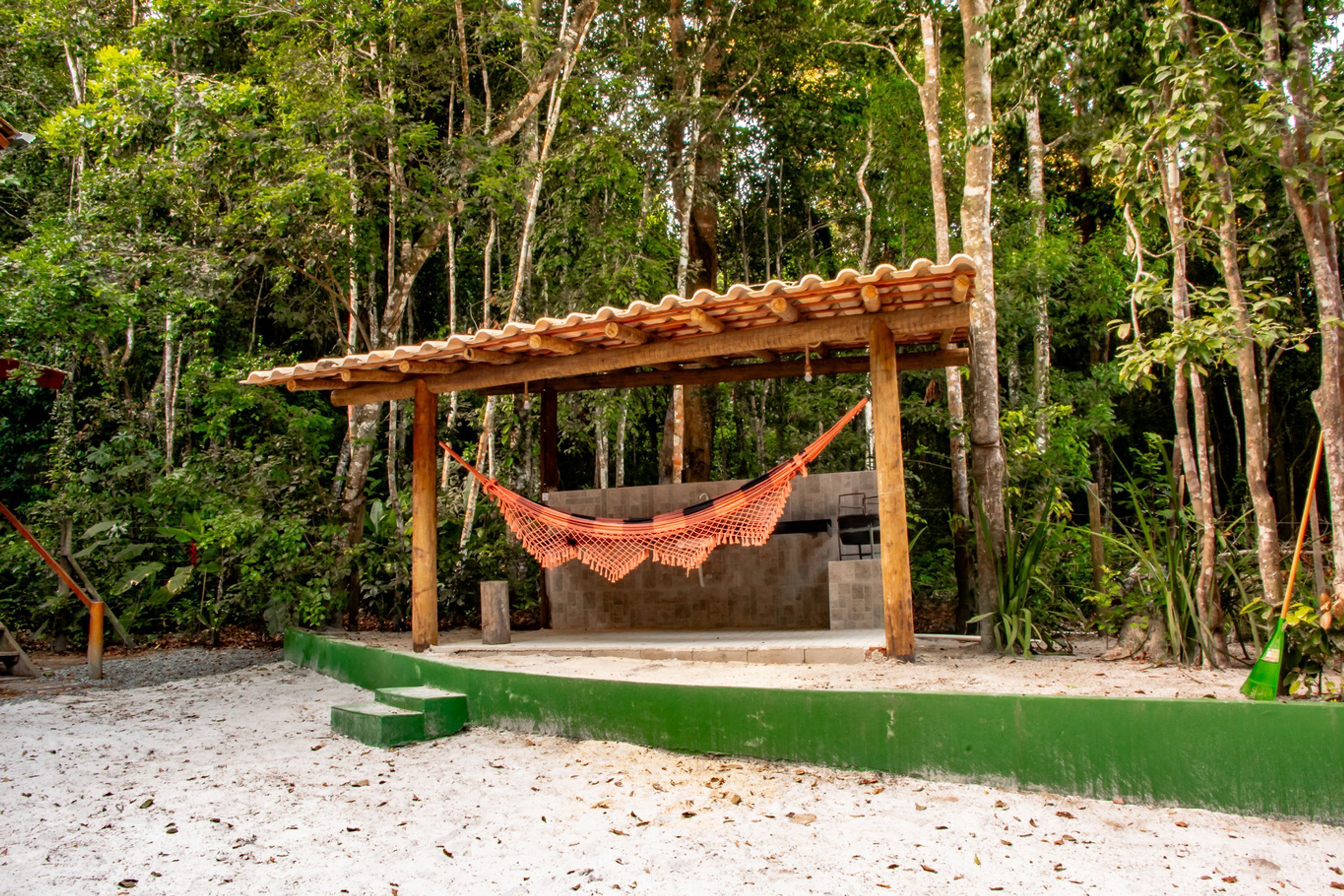A hammock under a wooden pergola in a forested area with a sandy ground.