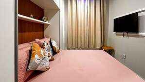 A bedroom with a bed, pillows, a TV, a shelf, and a window with curtains. - Timo Studio c Wifi a 190m da Praia do Leme RJ (Rio de Janeiro)