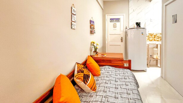 A bedroom with a bed, pillows, a small table with a vase of flowers, a refrigerator, and a door with a sign. - Timo Studio c Wifi a 190m da Praia do Leme RJ (Rio de Janeiro)