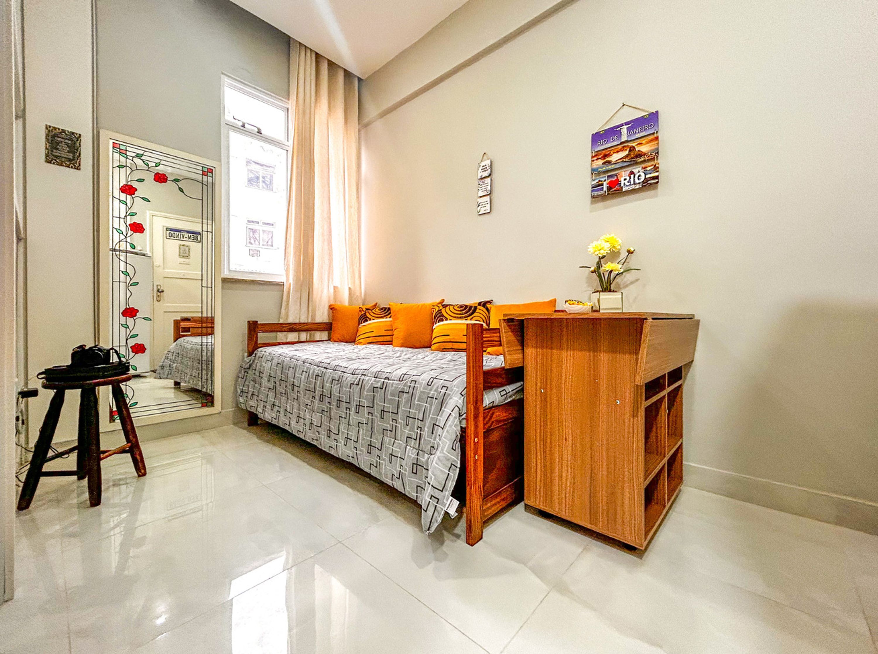 A bedroom with a bed, a wooden desk, a chair, a mirror, and a window with curtains.