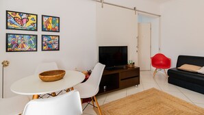 A modern living room with a TV, white chairs, a dining table, and colorful artwork on the wall. - Timo Apto c Wifi a 700m da Praia do Tombo-guaruj (Guarujá)