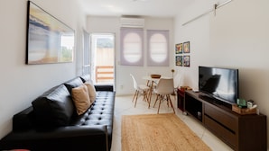 A modern living room with a black leather sofa, a flat-screen TV, a dining table with chairs, and a large window with blinds. - Timo Apto c Wifi a 700m da Praia do Tombo-guaruj (Guarujá)