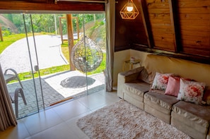 A living room with a sofa, a hanging chair, and a view through a glass door to an outdoor area. - Charmoso Chal no Campo com Vista Para a Natureza (Sao Pedro de Alcantara)