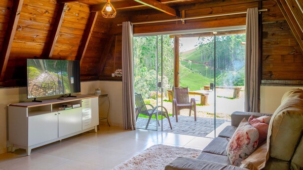 A cozy living room with a television, sofa, and a view of a garden through large glass doors. - Charmoso Chal no Campo com Vista Para a Natureza (Sao Pedro de Alcantara)