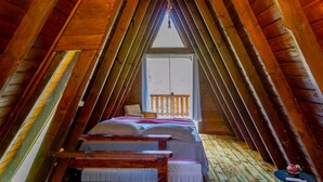 A cozy attic bedroom with a wooden sloped ceiling, a bed with a white and pink bedspread, and a small round table with a vase of flowers. - Charmoso Chal no Campo com Vista Para a Natureza (Sao Pedro de Alcantara)