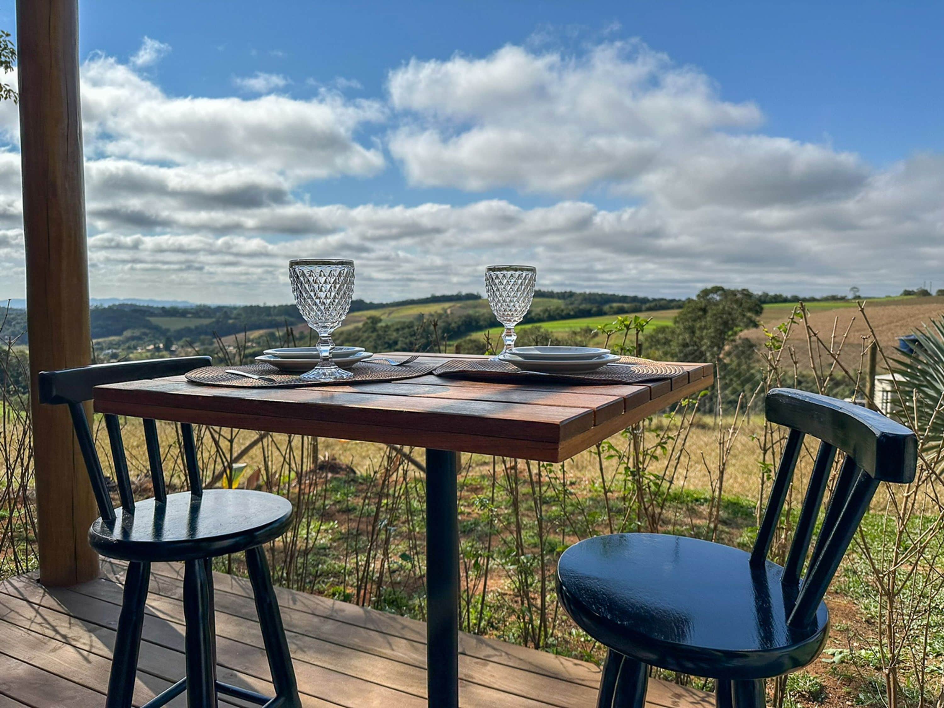 Uma mesa de madeira com duas cadeiras em um deck, com vista para uma paisagem rural com campos e um céu nublado.