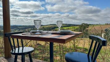 Uma mesa de madeira com duas cadeiras em um deck, com vista para uma paisagem rural com campos e um céu nublado.