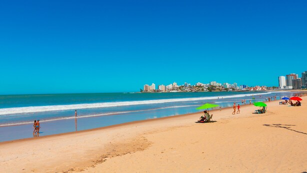 Una playa con cielo azul despejado, orilla arenosa y un horizonte urbano de fondo.