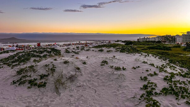 A coastal landscape with sand dunes, sparse vegetation, and a distant cityscape during sunset. - Incr vel Apto a 800m da Praia do Forte - Cabo Frio (Cabo Frio)