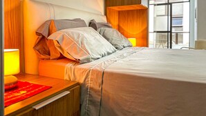 A modern bedroom with a wooden headboard, a bedside table with a lamp, and a view of the city through large windows. - Incr vel Apto a 800m da Praia do Forte - Cabo Frio (Cabo Frio)