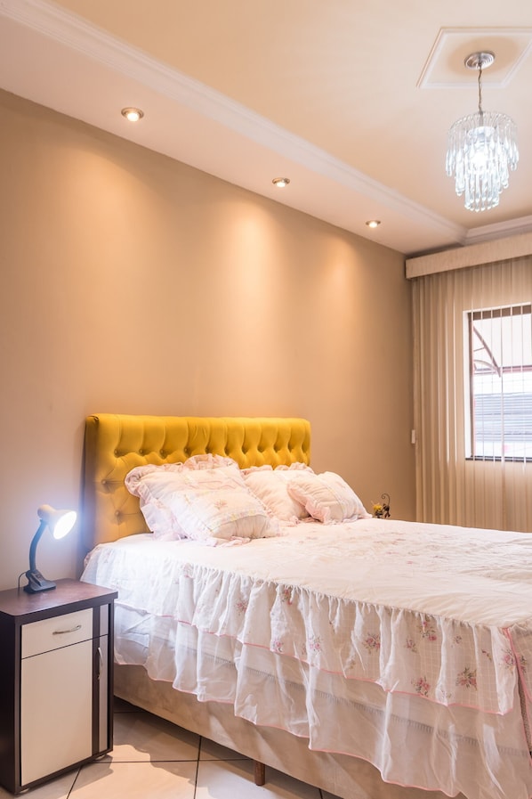 A bedroom with a yellow tufted headboard, a white bedspread with floral patterns, a bedside lamp, and a chandelier.