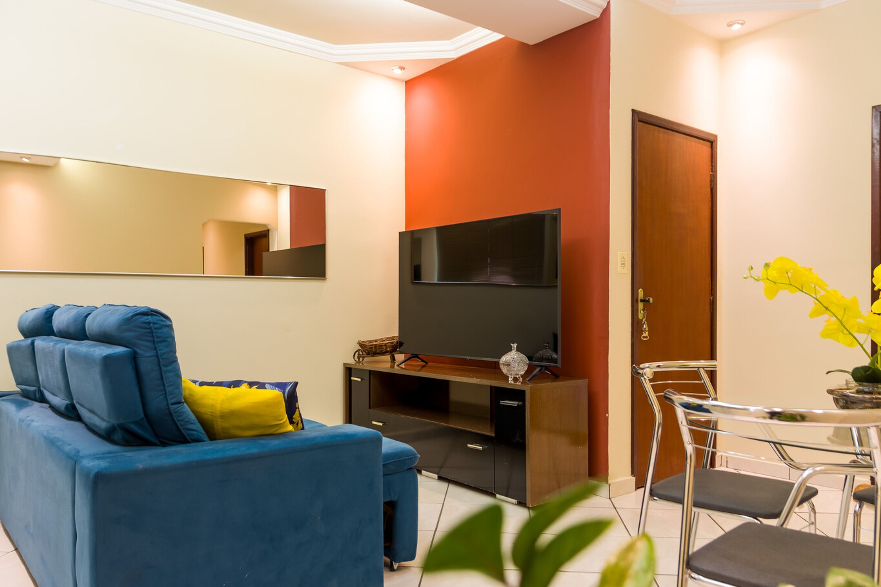 A modern living room with a blue sofa, a flat-screen TV, a dining table with chairs, and a wooden door.