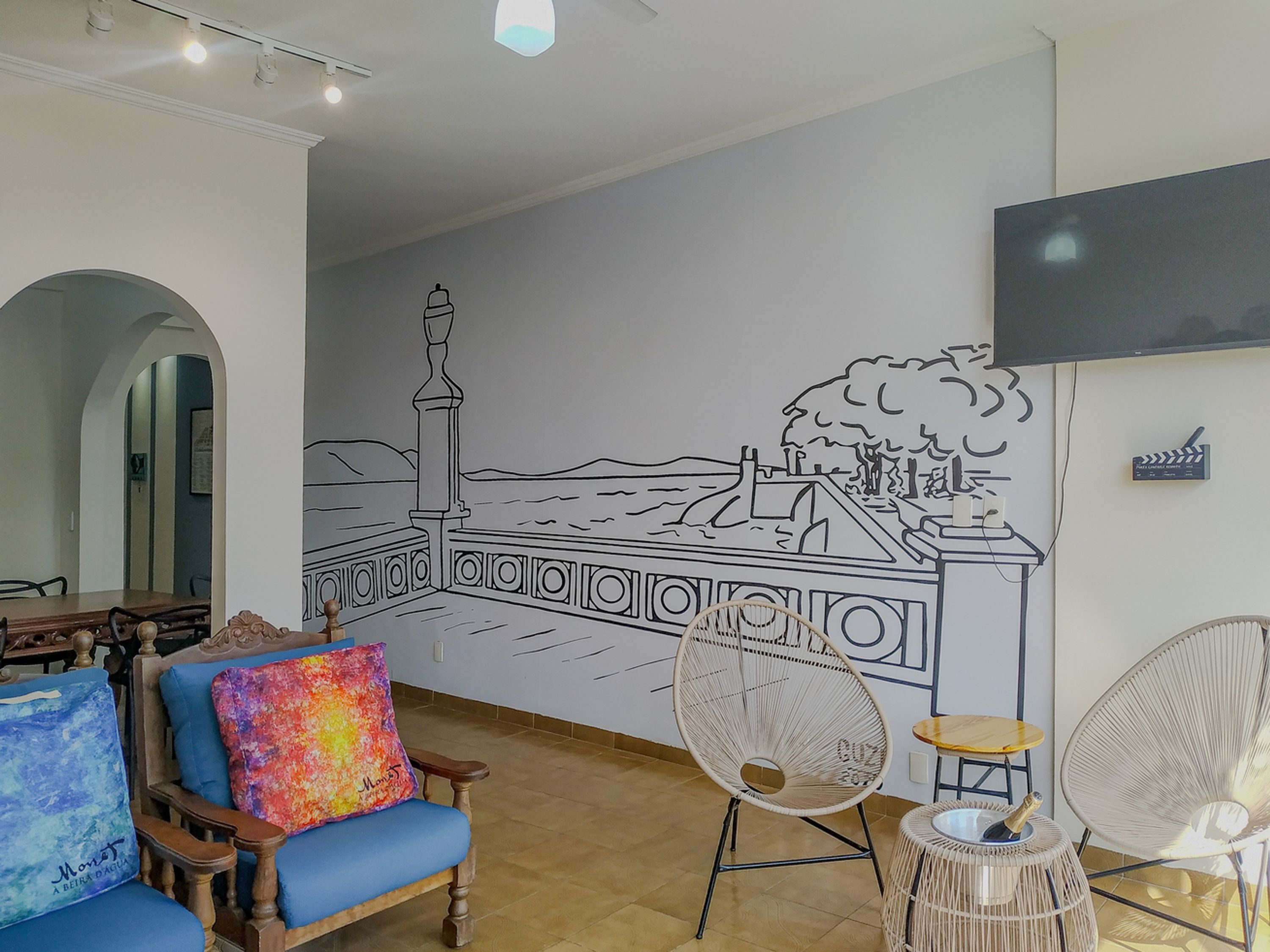 A room with a mural of a coastal scene, a wooden dining table, blue and colorful chairs, and two wicker chairs with a small table.