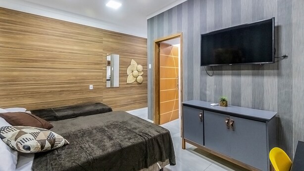 A modern bedroom with a wooden wall, a flat-screen TV, a cabinet, and a bed with a dark cover. - Loft pr x do Aeroporto no Centro e com Wi-fi (Rio de Janeiro)