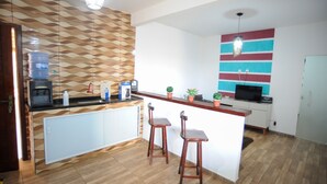 A compact kitchen with a water dispenser, coffee maker, and a small TV. There are two wooden stools and a countertop with potted plants. - Casa Beira-mar com Piscina em Saquarema RJ (Saquarema)