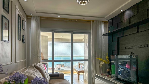 A living room with a sofa, a television, and a view of the sea through large windows. - Timo Apto de Frente Para Praia do Boqueir o SP (Praia Grande)
