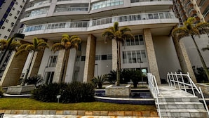 A tall, modern building with a curved facade, multiple balconies, and a well-maintained garden area with palm trees. - Timo Apto de Frente Para Praia do Boqueir o SP (Praia Grande)