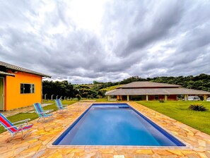 A swimming pool with stone tiles, two lounge chairs, and a small building with a tiled roof. - Tima ch Cara c Wi-fi e Churrasq em Socorro SP (Socorro)