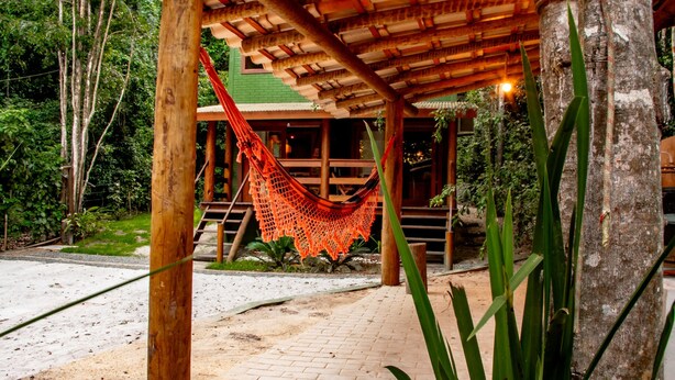 Estructura de madera con una hamaca, rodeada de follaje y una zona pavimentada.