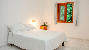 Una cama bien hecha con cubrecama y almohadas blancas, una mesita de noche de madera con una planta y una ventana con vistas a zonas verdes.