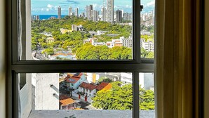 A window with a view of a cityscape, including buildings and greenery. - Apto Aconchegante c Wifi Ar-condicionado e Garagem (Salvador)