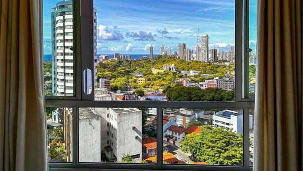 A cityscape through a window with curtains, featuring high-rise buildings, greenery, and a clear sky. - Apto Aconchegante c Wifi Ar-condicionado e Garagem (Salvador)