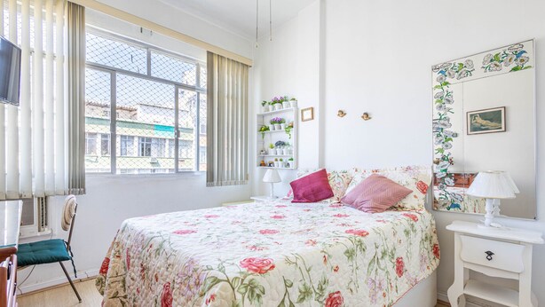 A bedroom with a floral bedspread, a bedside table with a lamp, a chair, and a window with curtains. - Timo Apto com Wi-fi a 350m da Praia de Copacabana (Rio de Janeiro)