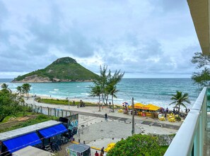 A beachside view with a mountain, a café, and a blue-roofed structure. - Flat a Beira mar no Recreio dos Bandeirantes RJ (Rio de Janeiro)