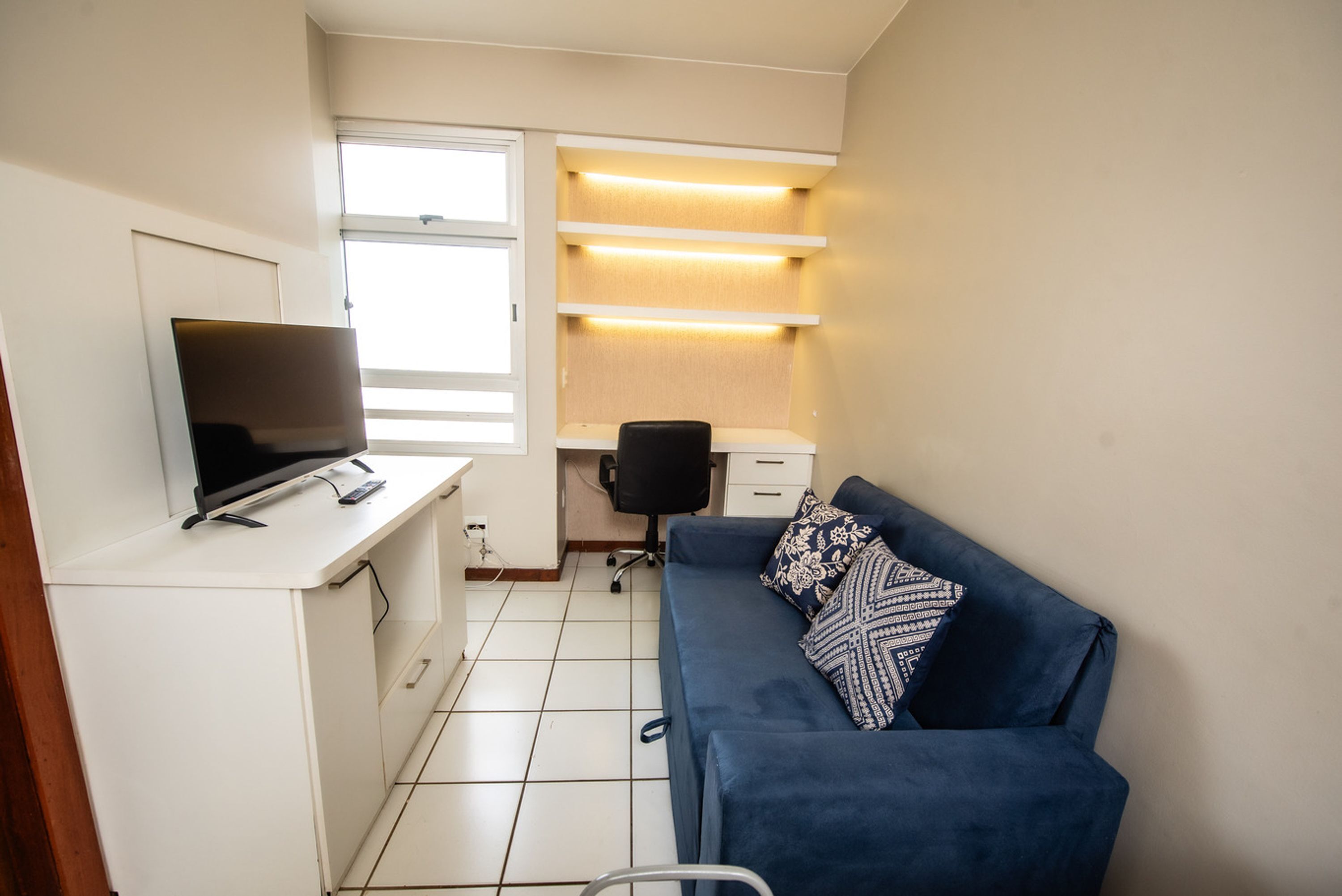 A compact room with a TV on a white cabinet, a blue sofa with patterned cushions, a desk with a chair, and a window with blinds.